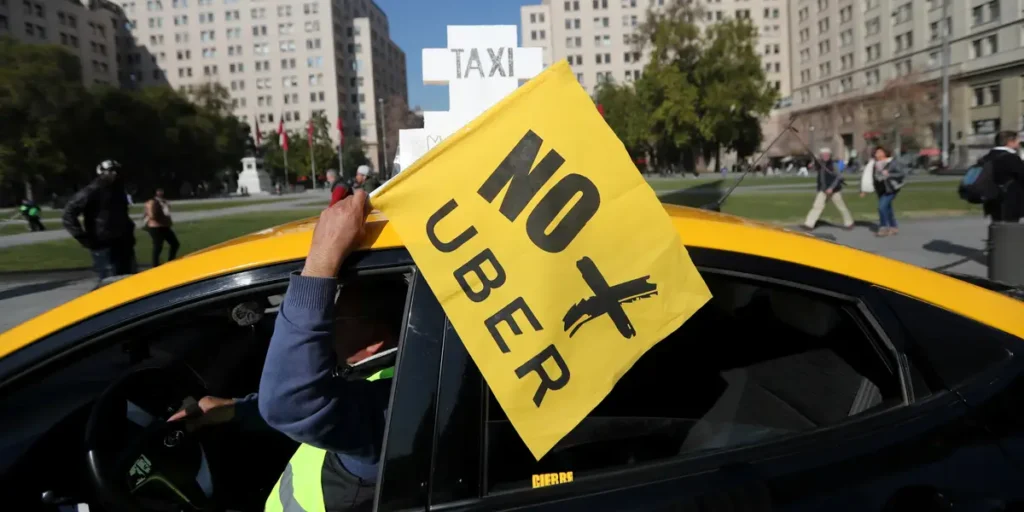 Por que empresas de transporte devem investir na comunicação em concessões públicas 5 Taxista dentro de um carro amarelo segura uma bandeira amarela com os dizeres “NO UBER” em letras pretas, expressando protesto contra o serviço de transporte por aplicativo. Ao fundo, há um parque urbano com pessoas caminhando e prédios altos ao redor, sugerindo que o protesto ocorre em uma área central de uma grande cidade.