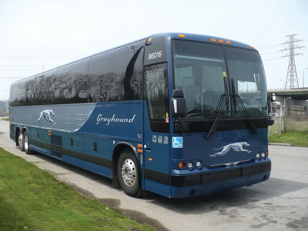 Por que empresas de transporte devem investir na comunicação em concessões públicas 6 Ônibus azul da empresa Greyhound estacionado em uma rua asfaltada, com o logotipo do galgo em movimento estampado na lateral e na frente do veículo. O ônibus é adaptado para acessibilidade, como indica o símbolo internacional ao lado da porta. Ao fundo, há uma cerca metálica, gramado e estruturas de viaduto e linhas de energia elétrica.