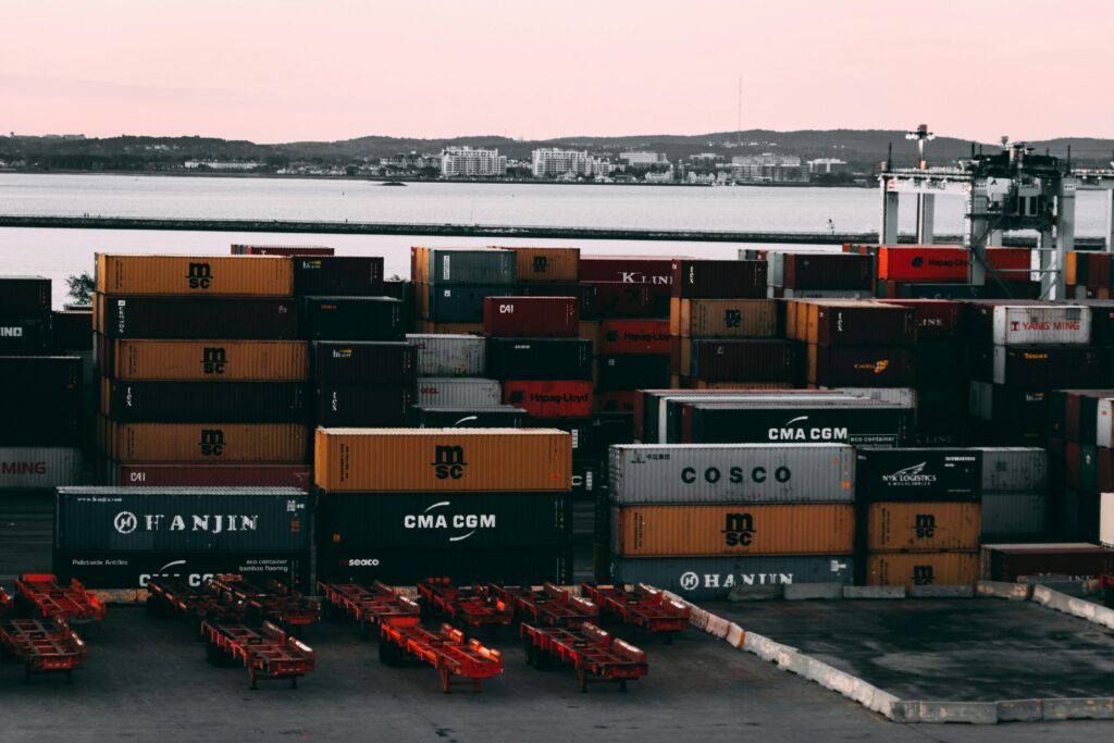 Como conseguir clientes para logística usando marketing digital 2 Pilha de contêineres de diversas companhias no porto à beira-mar, com empilhadeiras e estrutura portuária ao fundo.