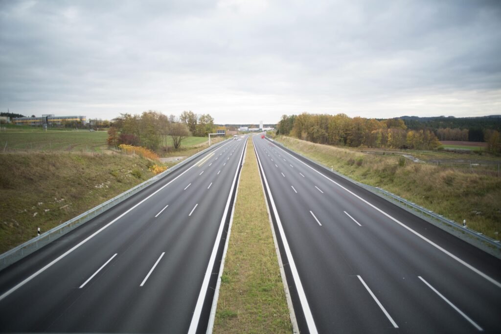 Como aplicar marketing industrial no setor de transportes 4 Vista aérea de uma rodovia vazia de múltiplas pistas com divisor central gramado, cercada por campos verdes e áreas florestais com árvores em tons outonais. O céu está nublado e há algumas construções visíveis ao fundo da paisagem rural.