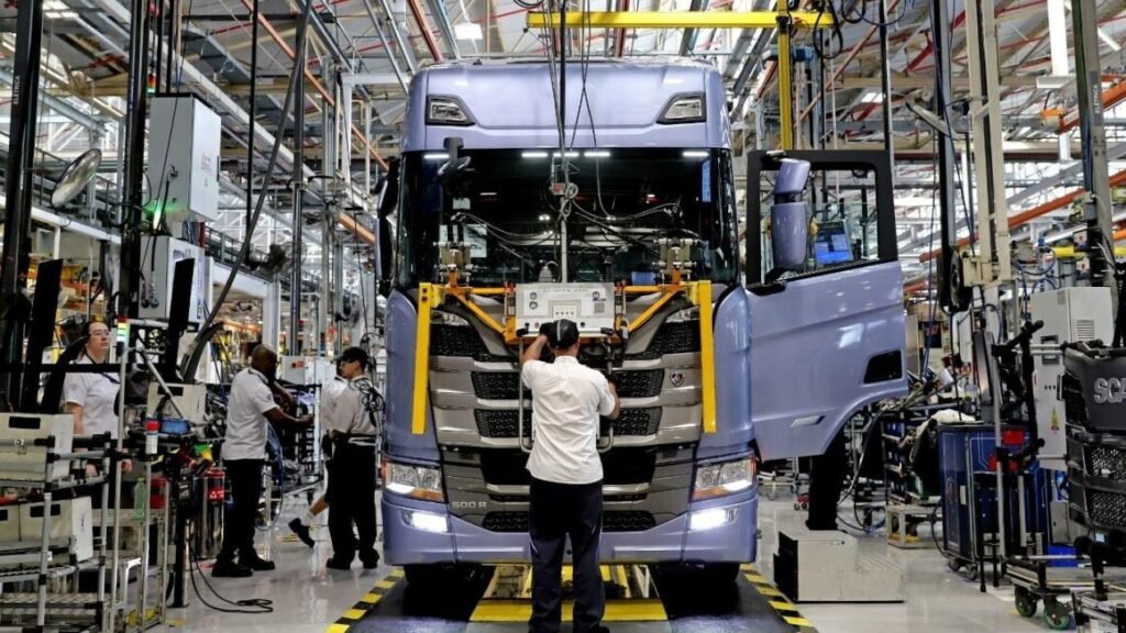 Como aplicar marketing industrial no setor de transportes 3 Trabalhadores em uniformes brancos montando um caminhão Scania azul e branco em uma linha de produção industrial. O veículo está suspenso por equipamentos amarelos em uma fábrica moderna com estruturas metálicas, iluminação industrial e diversos equipamentos de montagem ao fundo.