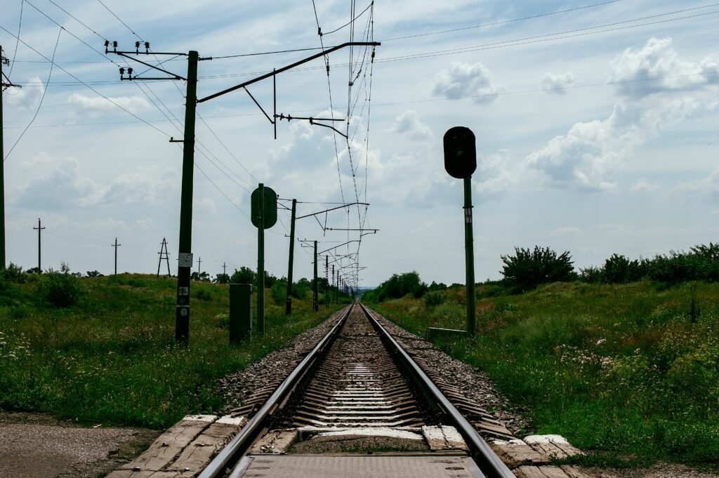 Quais são os tipos de sinalização ferroviária existentes? 4 Vista de trilhos ferroviários em área rural durante o dia, com postes de sinalização verdes, cabos de energia elétrica suspensos e vegetação verdejante nas laterais sob um céu parcialmente nublado.