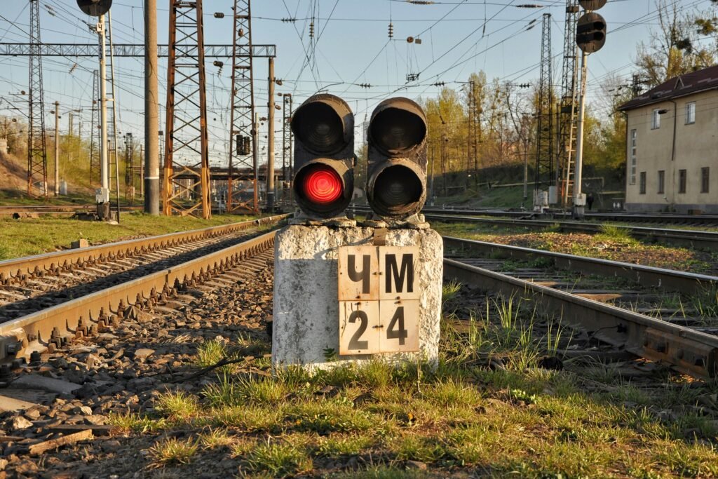 Quais são os tipos de sinalização ferroviária existentes? 3 Semáforo ferroviário com luz vermelha acesa e placa marcadora "ЧМ 2-4" posicionado entre trilhos duplos, com infraestrutura ferroviária complexa, torres de energia e edifício ao fundo em ambiente industrial.