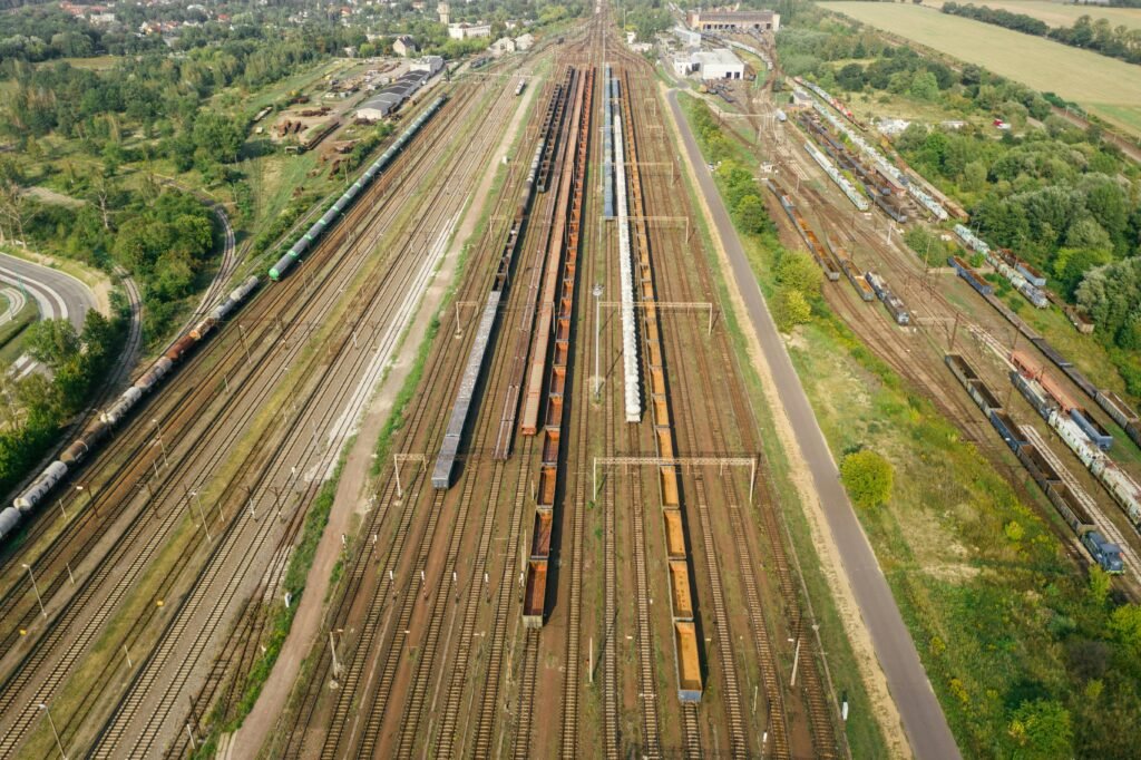 Como posicionar sua empresa nos projetos ferroviários brasileiros 3 Vista aérea de um amplo pátio de triagem ferroviária. A imagem destaca a escala logística do local, com dezenas de linhas férreas paralelas ocupadas por extensas composições de vagões de carga de diversos tipos, incluindo vagões-tanque e gôndolas. O complexo de infraestrutura é cercado por áreas verdes, estradas e algumas construções ao fundo.