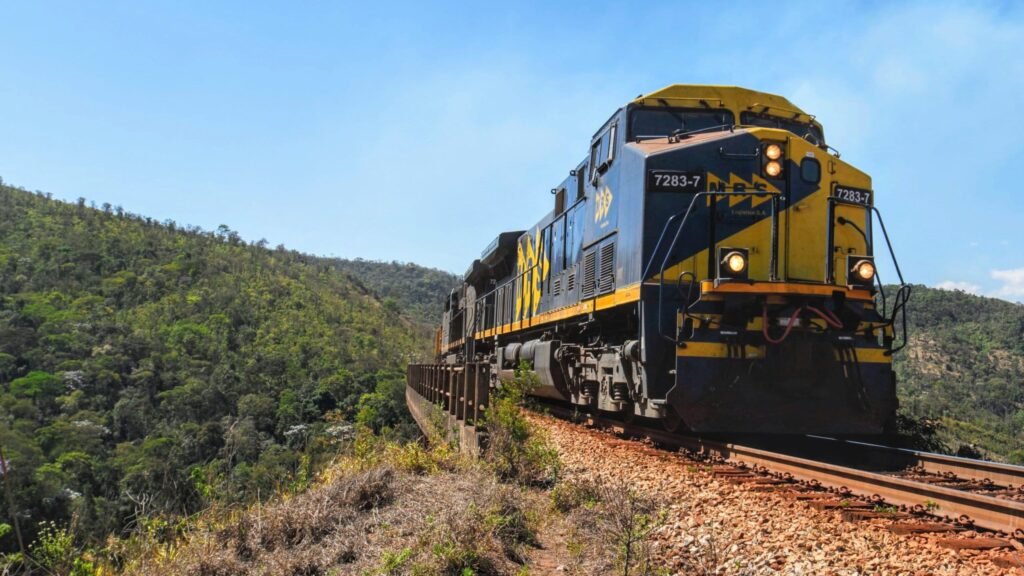 Como aplicar marketing de conteúdo para empresas de transportes 4 Locomotiva amarela e azul da MRS Logística em movimento sobre trilhos, cercada por montanhas e densa vegetação verde. A ferrovia é um modal estratégico no marketing de conteúdo para empresas de transportes.