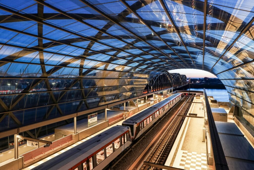 Estação ferroviária moderna com cobertura de vidro e metal, plataformas iluminadas ao entardecer e trens parados próximos a um rio.